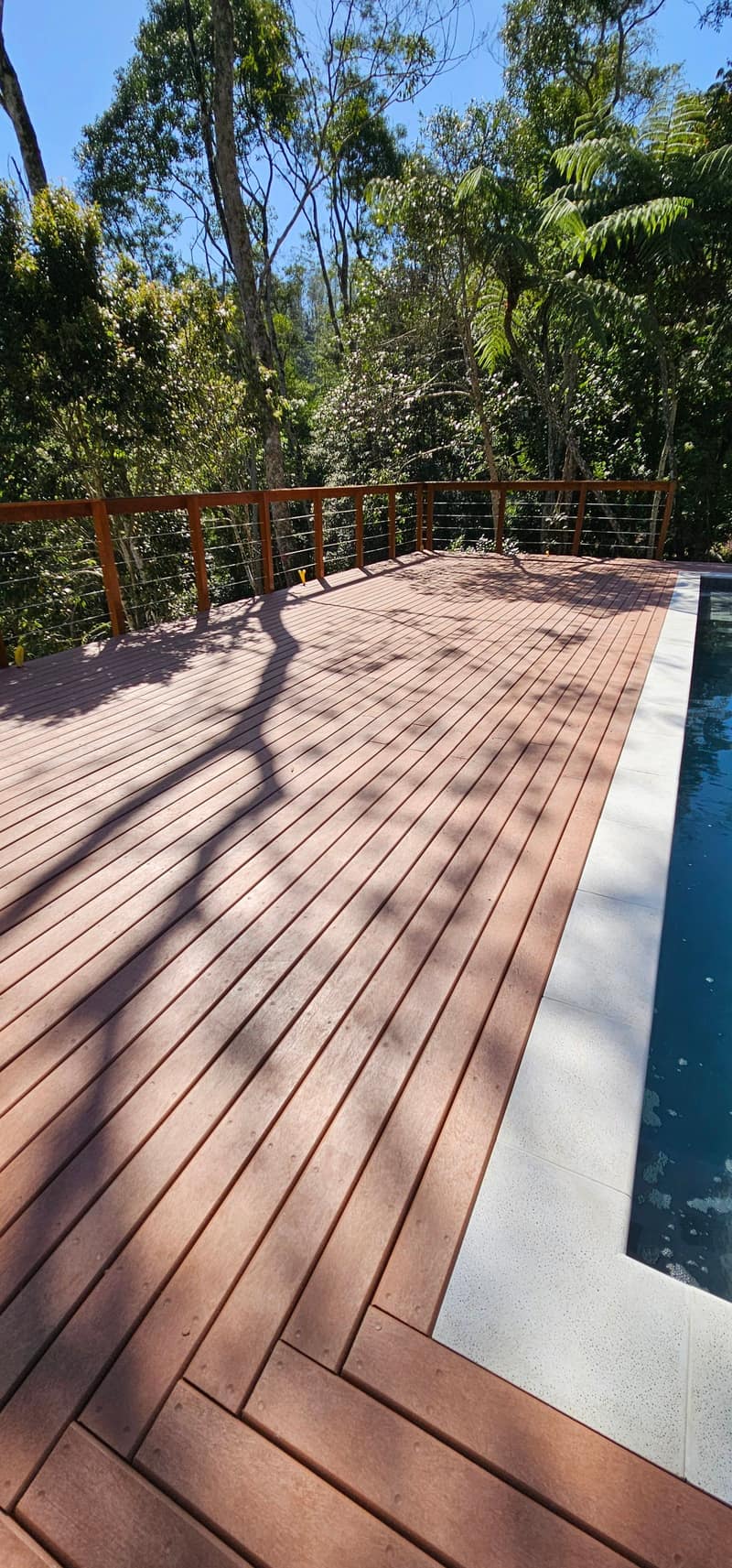 Deck longo de EcoDeck Supremo ao lado da piscina, cercado de mata